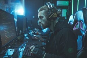Young man wearing a headset sits in a blue-lit gaming setup, typing at a keyboard with code visible on the monitor in front of him.”