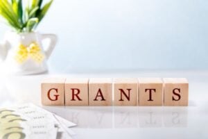 Wooden letter blocks spell 'GRANTS' on a reflective white surface, with a plant in a white pot in the blurred background.
