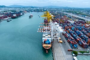 A bustling port with stacked shipping containers, large cranes, and a container ship docked at a quay along a wide waterway