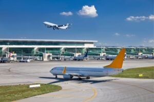 Airport - Funds for Companies A white airplane with a yellow tail taxis on the runway while another plane climbs in the blue sky near a modern terminal.