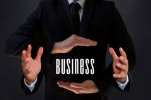 A person in a dark suit forms a protective circle with their hands around the word BUSINESS, symbolizing safeguarding or managing a company.