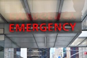 Red neon 'EMERGENCY' sign on a building overhang with glass panels and a city street in the background.