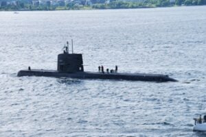 Dark submarine partially above water with several crew members on its deck at sea near a distant shoreline behind it.