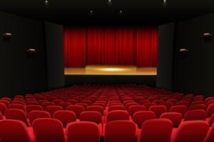 Front-to-back view of a dark theater with red velvet seats facing a lit stage behind closed red curtains.